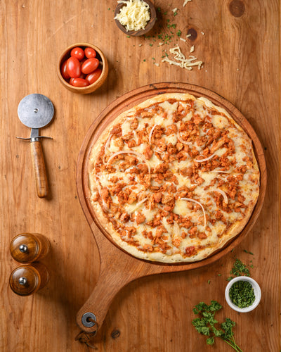 Wooden pizza peel with a pizza on a wooden table, surrounded by ingredients and tools.